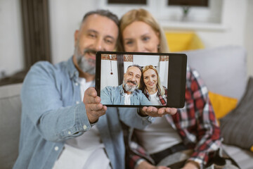 Close up of digital tablet screen with married couple that sitting closely on cozy sofa. Caucasian family taking selfie during free time at home.