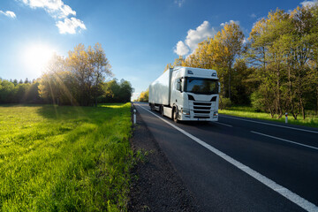 Wall Mural - white truck driving on the asphalt road between the forest and the meadow in springtime landscape at