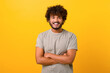 canvas print picture - Indian youngster looking at camera standing with arms crossed isolated on yellow background. Eastern male student with curly hair in casual t-shirt with folded hands