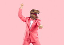 Funny Confident Man Wearing Rubber Dinosaur Mask Dancing At Show Or Event. Happy Crazy T Rex Guy In Funky Pink Party Suit With Bow Tie Doing Gangnam Style Moves Isolated On Pink Colour Background