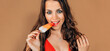 portrait of attractive brunette young woman with blue eyes and red lips biting popsicle looking at camera - copy space on  brown background