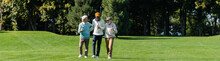 Senior Multiethnic Men Walking With Golf Clubs On Green Field Near Trees, Banner.