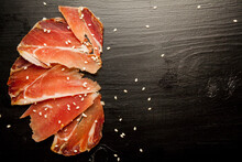 Sliced Jamon And Sesame Seeds On A Blackwood Background. Parma Ham, Hamon On A Black Background With Sesame Seeds With Space For A Textual Top View. Jamon Serrano, Iberico. Traditional Spanish Ham 
