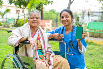 doctor showing green screen mobile phone wile patient sitting on wheelchair at park - concept of technology, healthcare app promotion and advertisement.