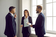 © Studio Romantic - Team of happy business people having a discussion. Group of three coworkers standing together by the window and talking. Smiling man and woman having a conversation with a more experienced colleague