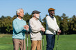 © LIGHTFIELD STUDIOS - senior man in flat cap near interracial friends standing with golf clubs.