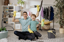 A Cheerful Man Holds Son In Lap As They Sit On The Laundry Room Floor. The Guys Are Cleaning The Bathroom Holding A Mop And A Spray Bottle With Liquid In Their Hands.