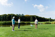 © LIGHTFIELD STUDIOS - back view of senior multiethnic friends with golf clubs standing on green lawn near pond.