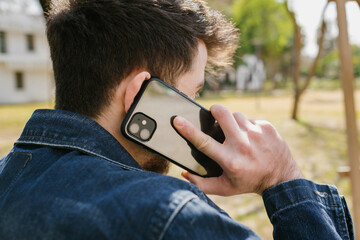 Wall Mural - Young man wearing blue jeans jacket from behind with mobile phone. Standing a city park while talking phone. Hidden face using mobile cell phone talking with victim request ransom.