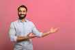 © Vadim Pastuh - Handsome Indian guy points palms at empty copy space isolated on pink background. Cheerful multiracial bearded man in casual jeans shirt presenting novelty, advertising concept
