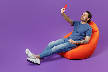 Full body young smiling man 20s wear basic blue t-shirt sit in bag chair doing selfie shot on mobile cell phone post photo on social network isolated on plain purple color background studio portrait