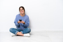 Young Caucasian Woman Sitting On The Floor Isolated On White Background Sending A Message With The Mobile