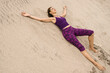 Latin woman wearing a purple leggings and top while lying on the sand at the beach spreading arms and legs and enjoying the sun and nature. Summertime and Relax concept. Copy space.