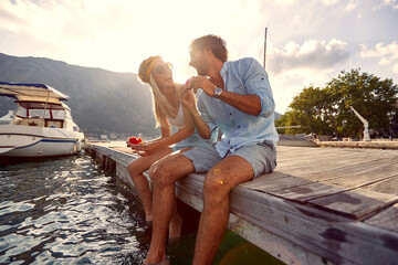 Lovely young couple sharing moments of bliss and eating watermelon sitting on wooden jetty by water. Summertime holiday by seaside. Togetherness, lifestyle, luxury concept.