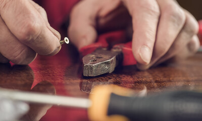 Older carpenter holding in hands metal screw and red pliers sitting at wooden desk