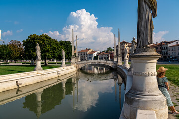 Woman in a dress and hat leaning against columns of a statue at Prato della Valle, square in the city of Padua, Veneto, Italy, Europe. Isola Memmia surrounded by canal bordered by two rings of statues