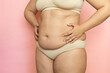 Clutch woman sagging belly closeup, folds on stomach, loose skin and cellulite, obesity. Naked overweight plus size girl on pink background in beige underwear. Concept of dieting and body control.