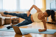 Fitness instructor doing side plank with leg raise while holding online exercise class over laptop at home.