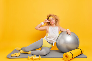 Sporty healthy woman makes peace gesture does sport exercises on fitness mat has workout at home with fitball and dumbbells isolated over yellow background. Active lifestyle routine concept.