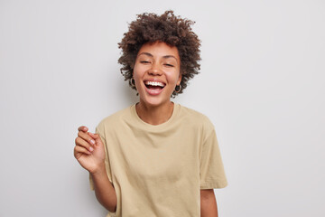 Wall Mural - Positive curly haired beautiful woman laughs happily has carefree expression keeps mouth opened wears casual beige t shirt isolated over white studio background. Sincere human emotions concept