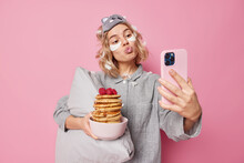 Lovely Woman Pouts Lips And Poses At Front Camera Of Smartphone Poses With Delicious Pancakes For Breakfast Wears Slumber Suit Holds Soft Pillow Applies Beauty Patches Poses Against Pink Wall