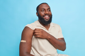 Bearded dark skinned adult man shows arm with plaster after coronavirus vaccination gets vaccine injection has glad expression poses against blue background. Immunity and health care concept