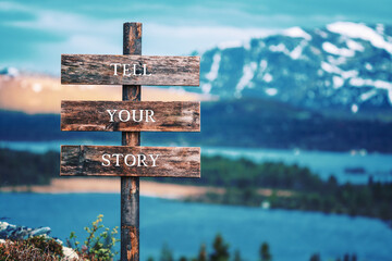 tell your story text quote written on wooden signpost outdoors in nature with lake and mountain scenery in the background. Moody feeling.