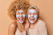 © WHstudio Leushin N - Overjoyed positive young women stand closely to each other keep mouthes opened apply beauty mask for skin care and moisturising dressed casually isolated over brown background. Wellness concept