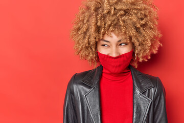 Wall Mural - Cheerful curly haired woman looks away happily wears turtleneck covers mouth with collar dressed in black leather jacket isolated over vivid red background empty space for your advertisement