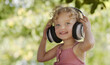 Happy smiling little baby girl listening to music wearing headphones, close-up portrait of funny kid with blue eyes and blond curly hair, laughing child looking at camera, outdoor in sunny summer day.