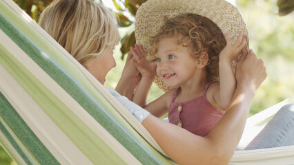 happy mothers day, smiling mom playing with her blue eyed little girl daughter child, plays with her by putting on a big straw hat on hammock in sunny green garden, spring and summer time concept