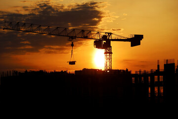 Wall Mural - Black silhouette of tower crane with cargo and workers on building scaffolding at sunrise. Housing construction, apartment block in city on dramatic sky background