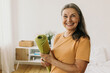© shurkin_son - Happy cheerful joyful mature 60-years-old sporty woman standing against bed at her apartment with rolled green mat in hands, waiting with anticipation beginning of daily physical workout