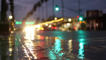 Cars lights reflection on road in rainy weather. Rain drops on wet asphalt of city street in USA, water raindrops falling on sidewalk. Palm trees and rainfall, twilight dusk. Ocean Beach, California.