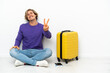 © luismolinero - Young blonde man with suitcase sitting on the floor smiling and showing victory sign
