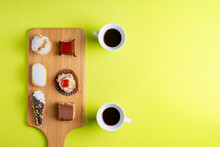 Various Cakes On A Wooden Board Next To Two Cups Of Coffee On A Green Background.Copy Space.