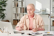 © Mediaphotos - Serious young bald woman in turtleneck sitting at desk with laptop and holding glass of water while taking pill in office