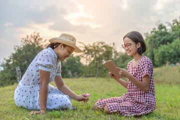 Canvas Print - Smile happy two teenage girls sitting on meadow playing and reading a book sunset background