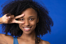 Smiling Woman Gesturing Peace Sign In Front Of Eye Against Blue Background