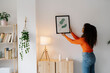 © Westend61 - Young woman with curly hair hanging picture frame on wall at home