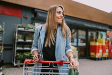 Smiling Woman With Smart Phone And Shopping Cart Standing In Front Of Store