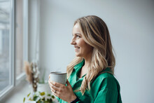 Smiling Woman Holding Coffee Cup Looking Out Through Window At Home