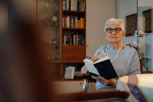 Senior Woman With Magnifying Glass And Book Sitting At Home