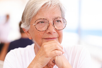 Wall Mural - Close up portrait of a beautiful senior caucasian woman with white hair and glasses sitting outdoors while looking thoughtfully away with her hands under her chin