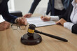 © Studio Romantic - Close up of a judge's gavel on a wooden table, and an experienced lawyer working with notary documents and giving legal advice to a group of clients in the background. Law services concept