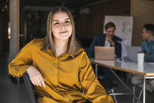 Confident Businesswoman Sitting On Chair In Office With Colleagues In Background