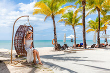 Young Attractive Woman In Straw Hat Rest In Rattan Wicker Cocoon Swing Chair By Beach In Luxury Beachfront Hotel Resort At Sunny Day, Enjoy Holiday Vacation. Sea, Blue Sky, Palm Trees On Background.