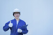 © Bond_JP - Portrait with copy space of female Asian woman in blue jumpsuit, white safety helmet and gloves, holding clipboard, smiling, putting thumb up. Engineer or worker, satisfaction and guarantee concept