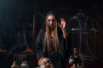 Wall Mural - Halloween concept. Witch portrait close up with dreadlocks dressed black hood standing dark room with cage on background