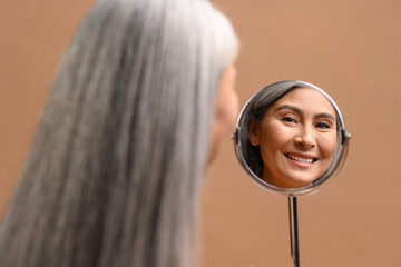 Poster - Delightful middle-aged 50s Asian woman with long gray hair and naked shoulders in reflection of round makeup mirror, isolated on brown background